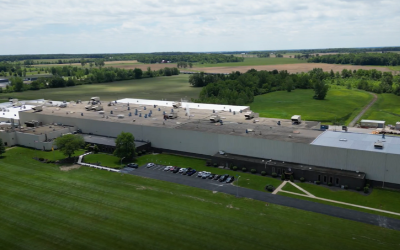 A large industrial building with a flat roof is surrounded by green fields and trees. Several cars are parked in front, and rural farmland stretches out in the background under a partly cloudy sky. - Anderson Smith Group
