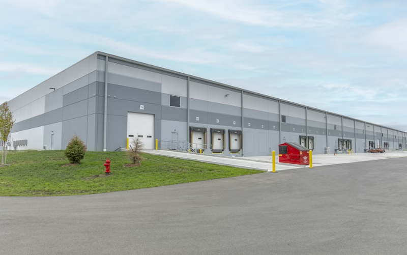 A large, modern warehouse with gray exterior walls, multiple loading docks, a red dumpster, and a few small trees on a grassy area near a paved road under a partly cloudy sky. - Anderson Smith Group
