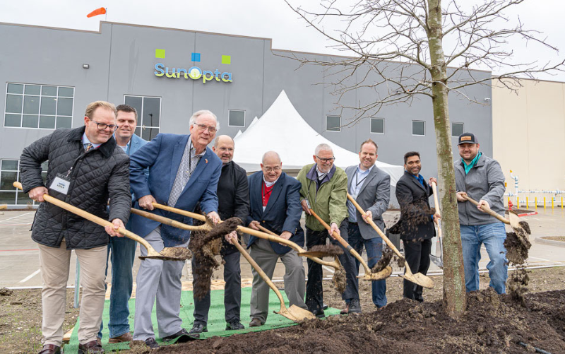 A group of nine people in business and casual attire smile as they plant a tree with shovels in front of a modern building with the SunOpta logo. A white tent is visible in the background. - Anderson Smith Group