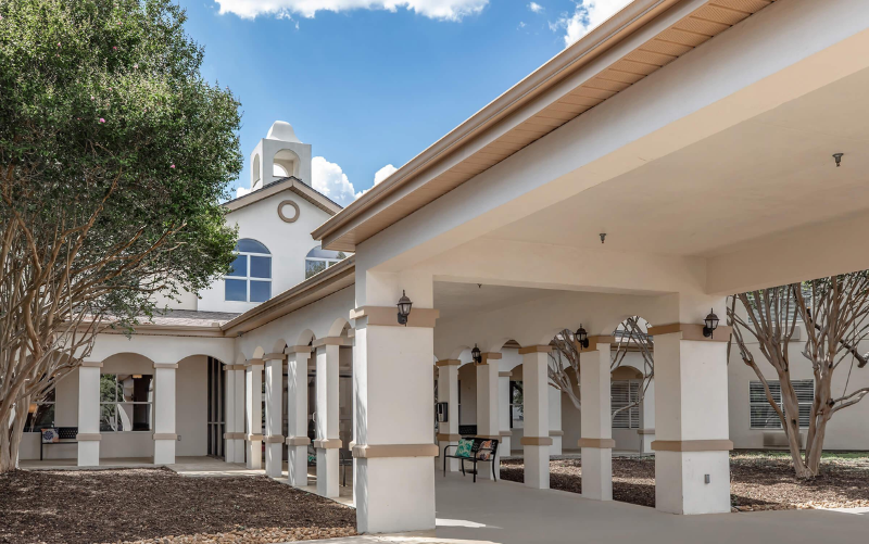 A large, white, single-story building with arches and columns, a covered walkway, and a small bell tower, surrounded by trees under a blue sky with clouds. - Anderson Smith Group