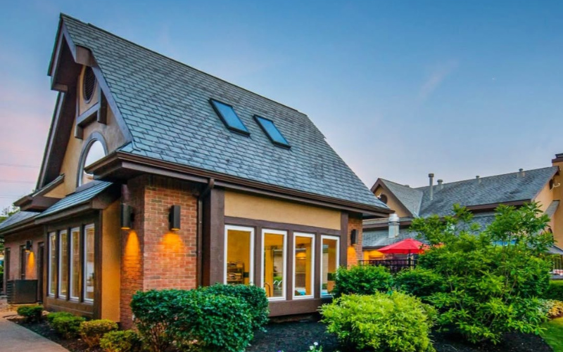 A charming house with a steep slate roof, brick and tan siding, large white-trimmed windows, skylights, and landscaped bushes, photographed at sunset with a blue sky. - Anderson Smith Group