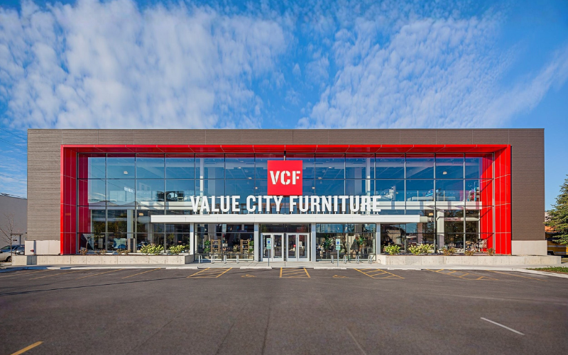 A large Value City Furniture store with a modern glass facade, red accents, and a sign reading VCF VALUE CITY FURNITURE. The building sits in front of an empty parking lot under a blue sky with scattered clouds. - Anderson Smith Group