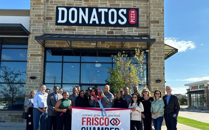 A group of people stand outside Donatos Pizza, holding a large Frisco Chamber banner and oversized scissors for a ribbon-cutting event. The restaurant sign is visible above the group on a sunny day. - Anderson Smith Group