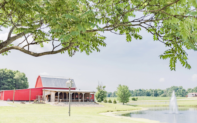A bright red barn sits beside a small pond with a water fountain, surrounded by green grass and trees under a clear sky. Tree branches frame the top of the image. - Anderson Smith Group