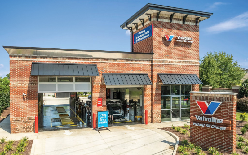 A Valvoline Instant Oil Change brick building with three service bays, an outdoor sign, and clear blue sky in the background. One vehicle is being serviced inside the center bay. - Anderson Smith Group