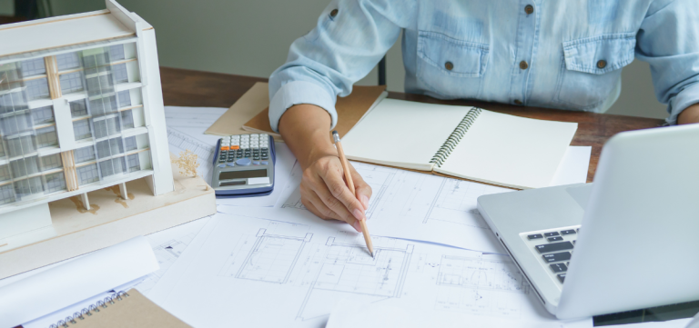 Person working at a desk with architectural blueprints, a model building, an open laptop, a notebook, and a calculator, appearing to make notes or edits on a drafting plan. - Anderson Smith Group